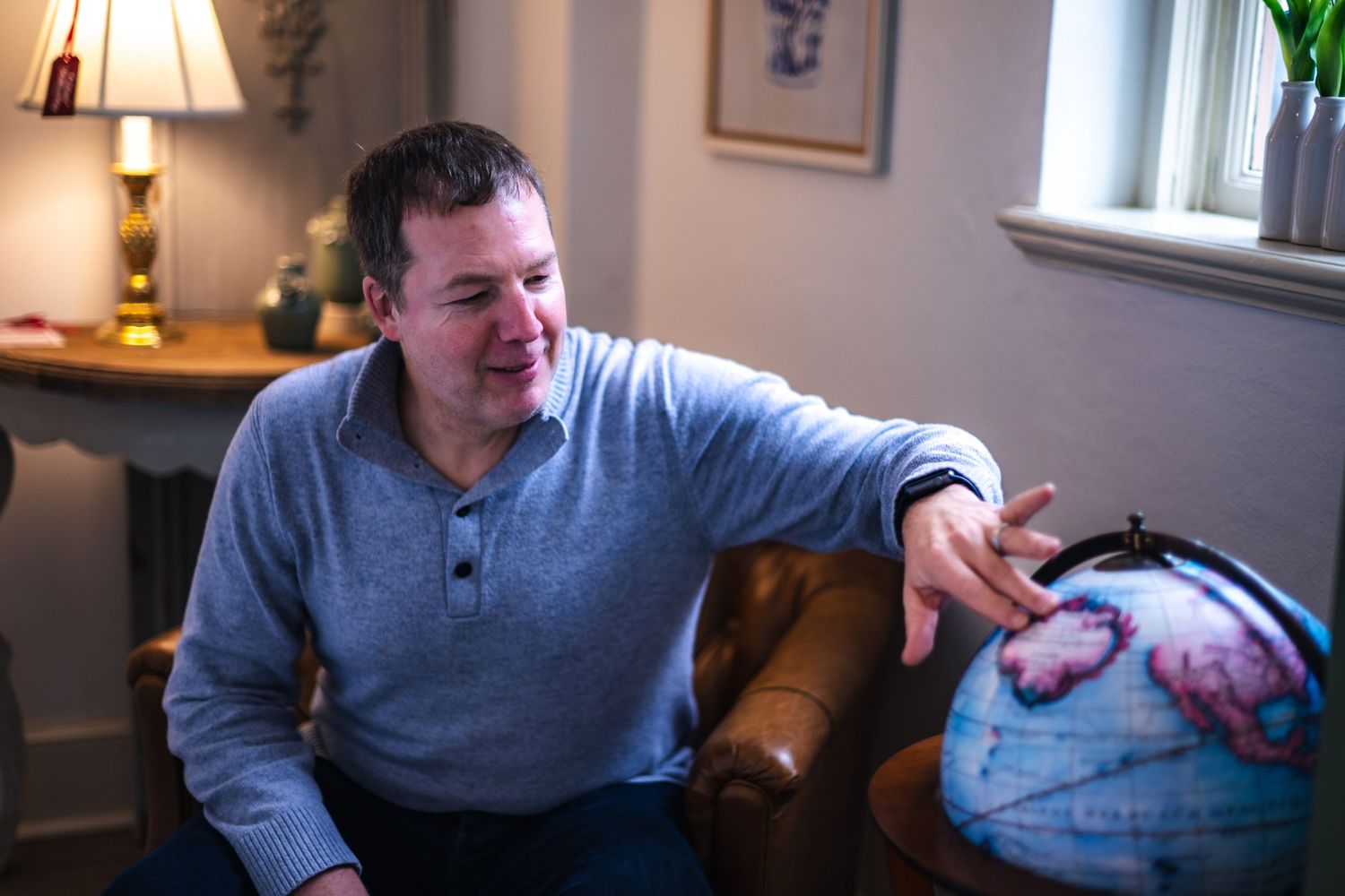 Someone in a gray henley shirt sits on a brown leather couch while pointing at a globe in a cozy living room setting.