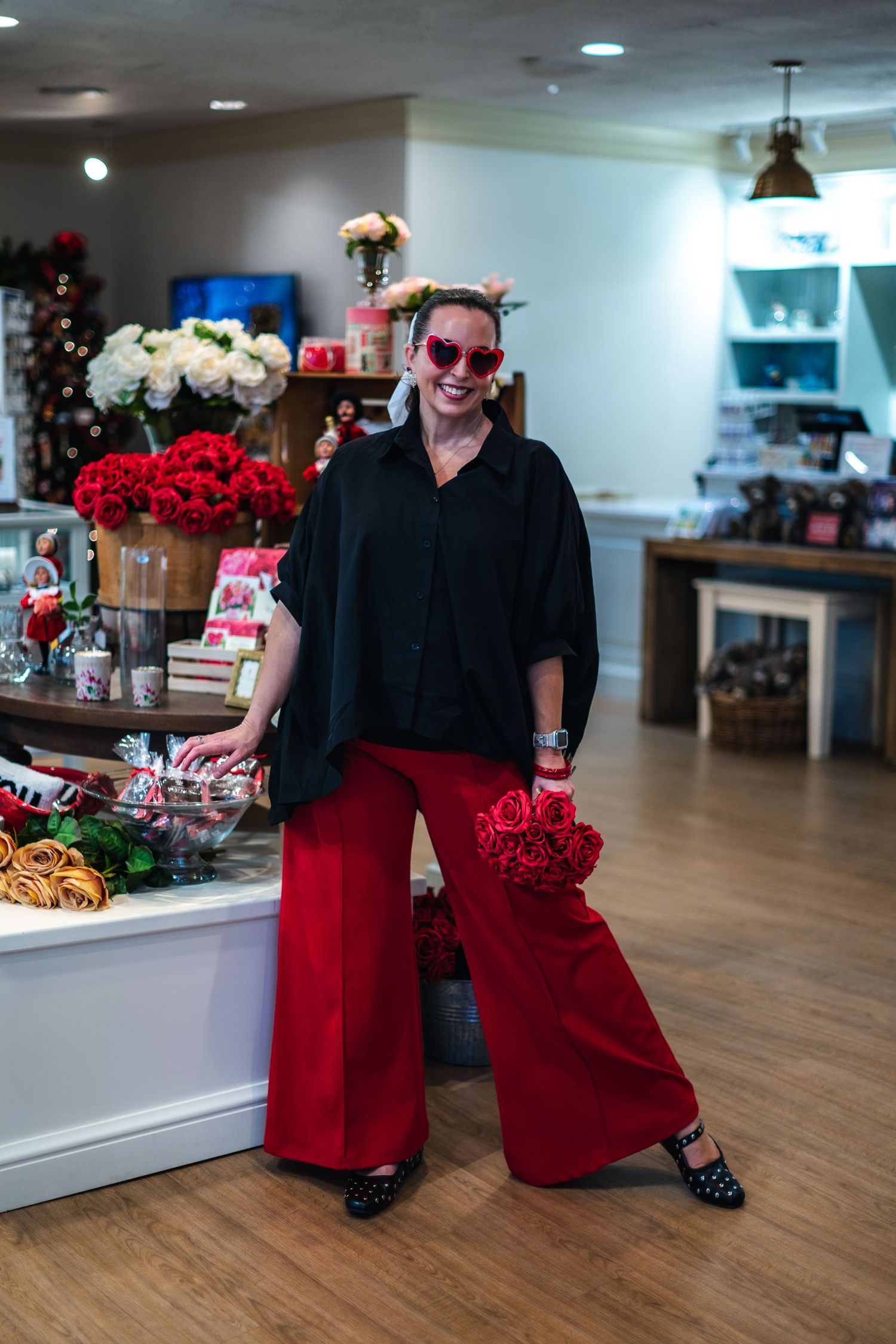 Someone wearing a black sweater and vibrant red palazzo pants poses in a retail space with holiday decor and floral displays.