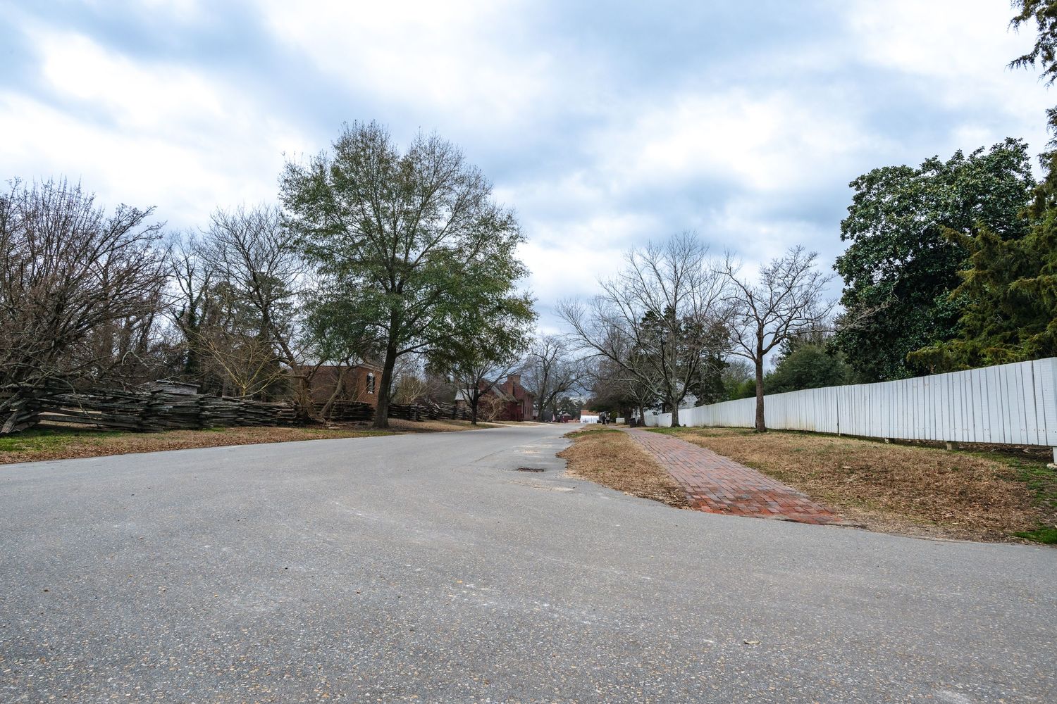 An empty curved road with a white picket fence and bare trees on a cloudy winter day.