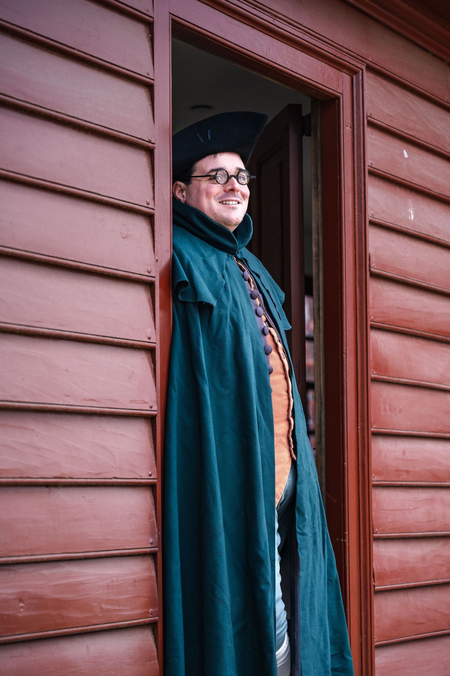 A smiling figure in a tri-corner hat and teal colonial-style cloak stands in a doorway of a rustic red building.