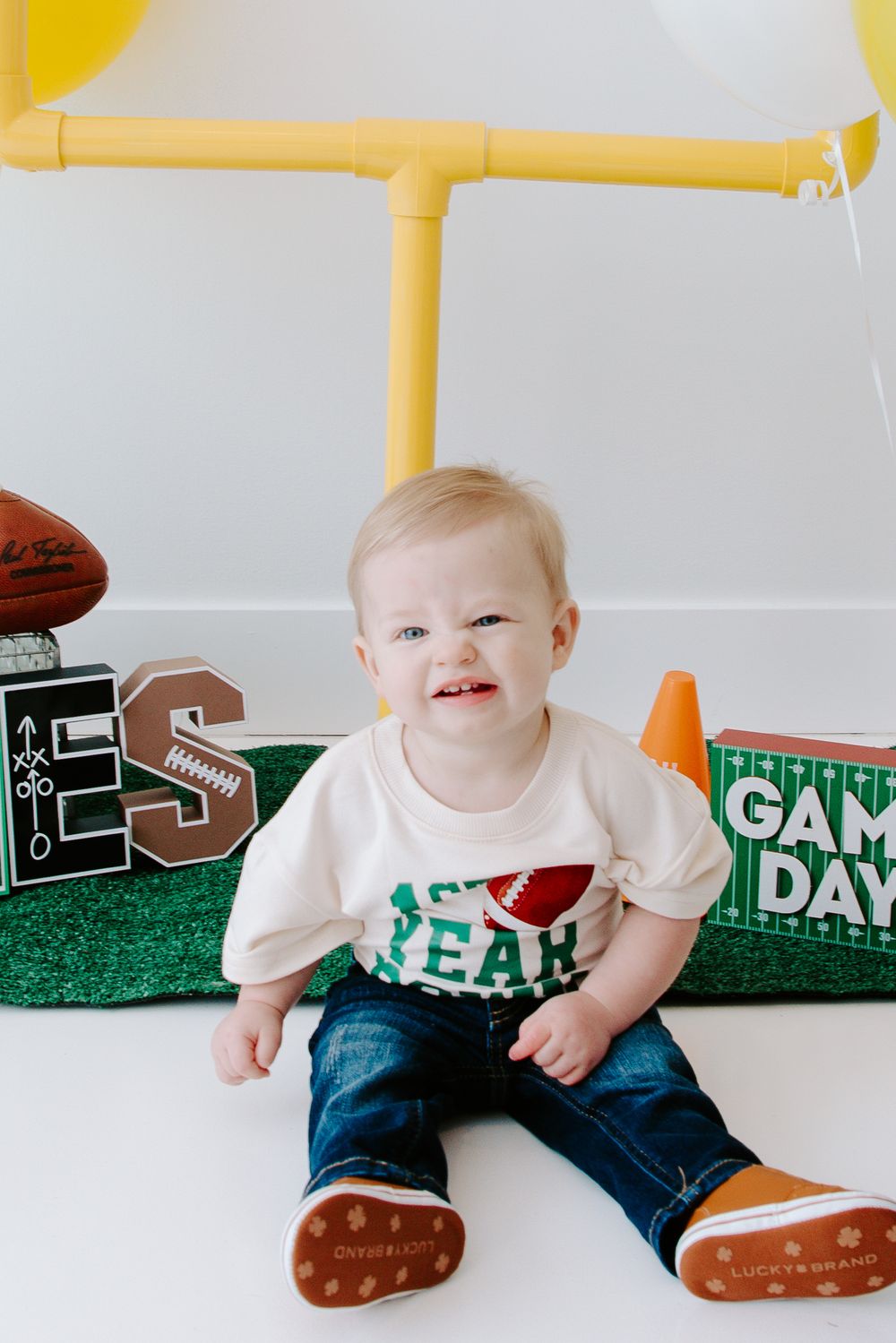 First birthday football party setup with letter decorations on artificial turf and yellow balloon backdrop.