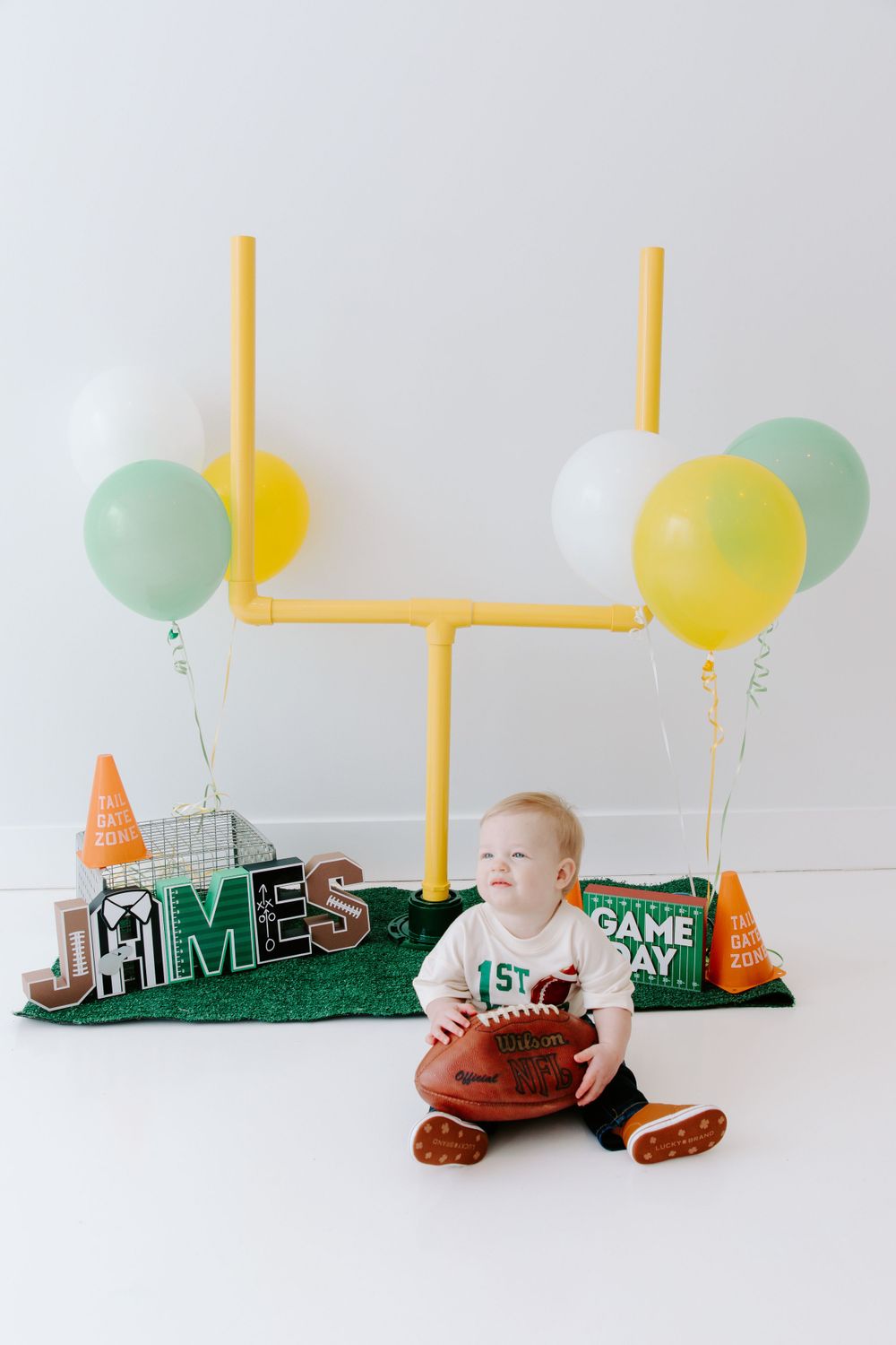 Football-themed baby photo setup with yellow goal post prop, green turf, balloons, and orange cones on white backdrop.