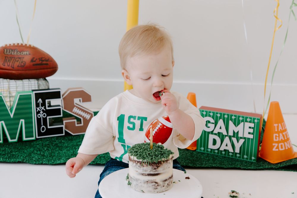 Baby in white shirt enjoying cake during first game day celebration with football decorations and party setup.