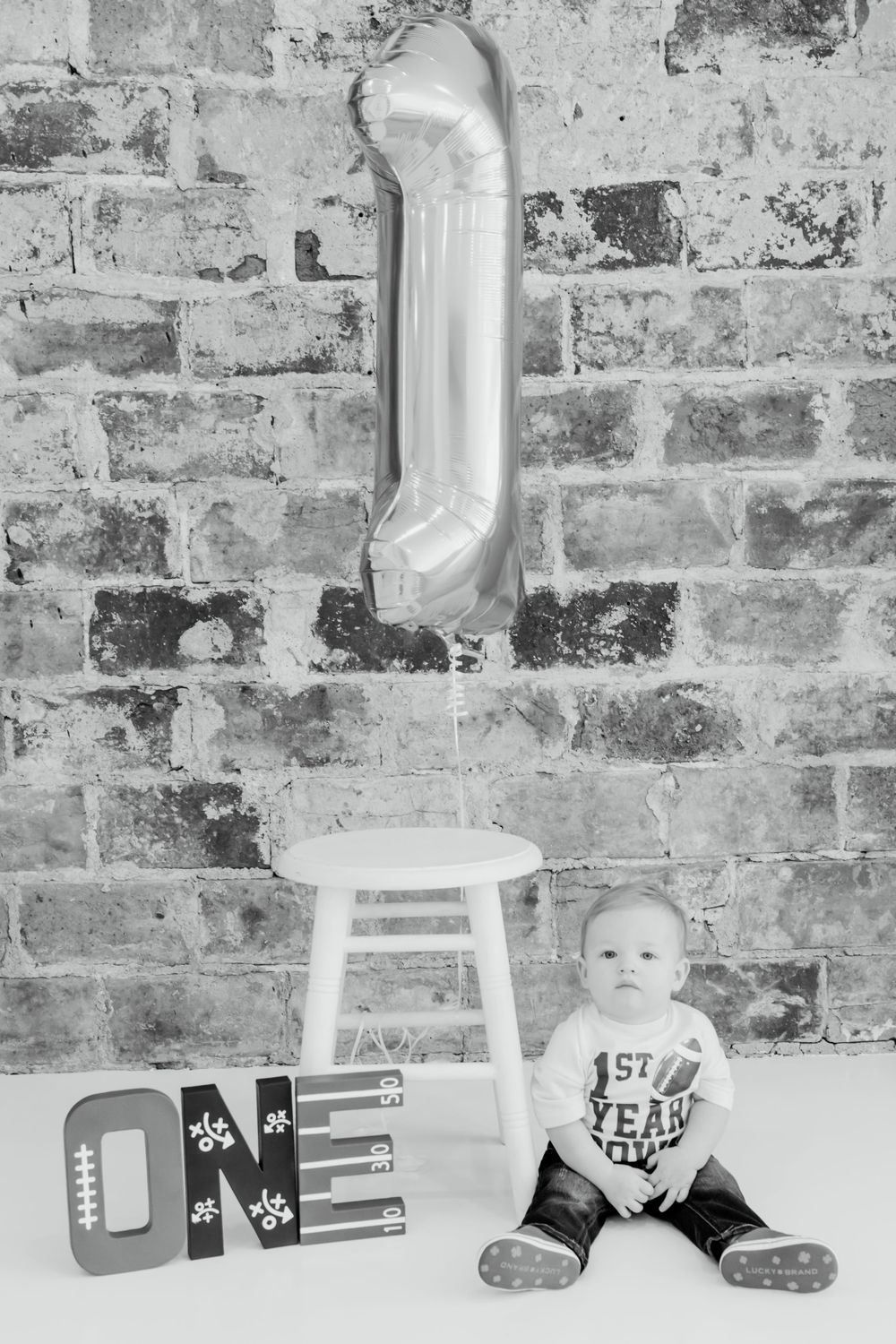 Black and white first birthday photo setup with silver balloon number one, wooden stool and brick wall backdrop.