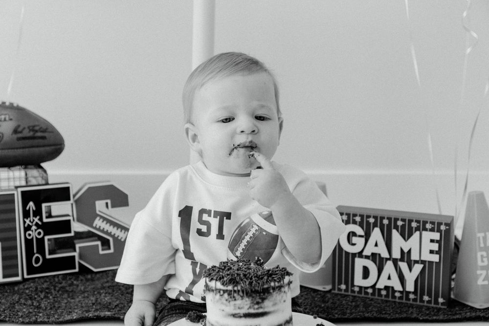 Black and white photo of baby eating cake at first game day celebration with football themed decor.