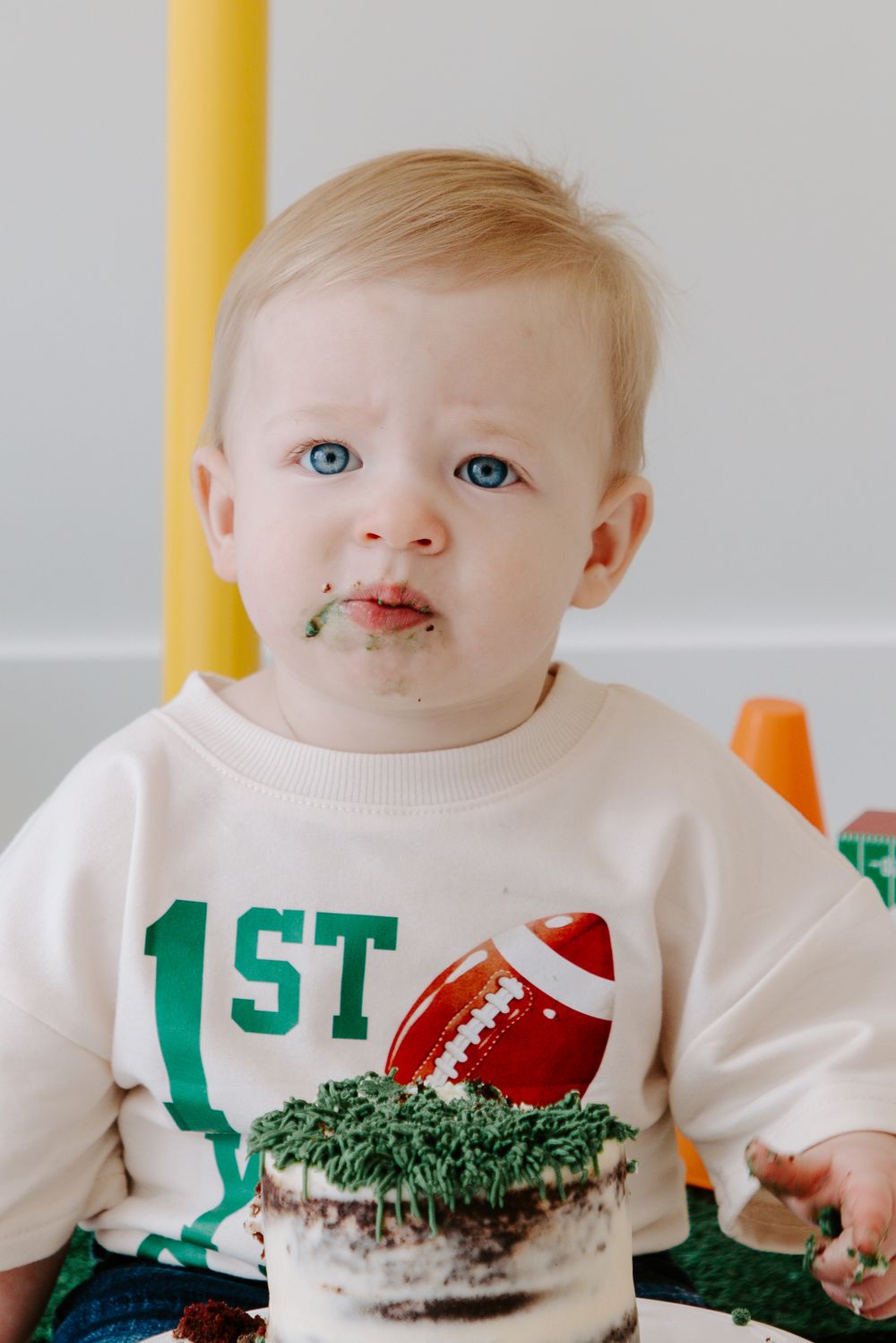 Baby wearing white football themed shirt enjoying first birthday cake smash.