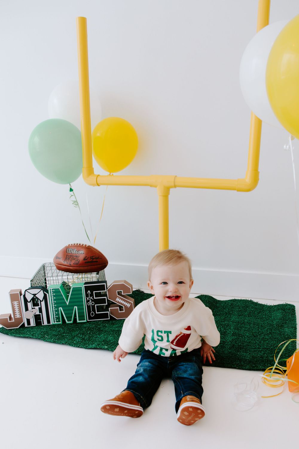 Baby sitting on turf with football decorations, yellow and mint balloons, and goal post backdrop.