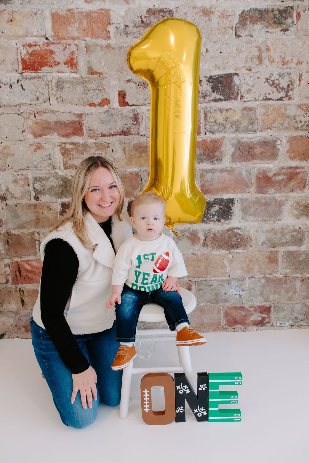 Football themed first birthday party setup with gold number 1 balloon against brick wall and decorative ONE letters.