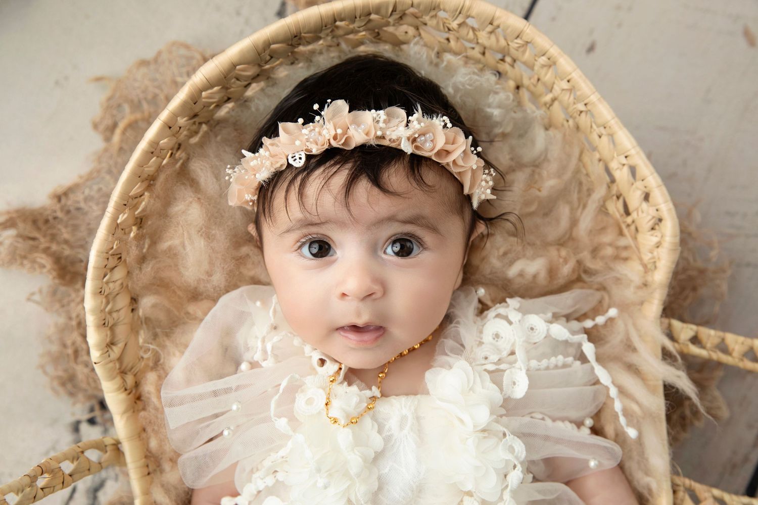 White lace dress and floral crown on infant in vintage wicker chair against beige background.