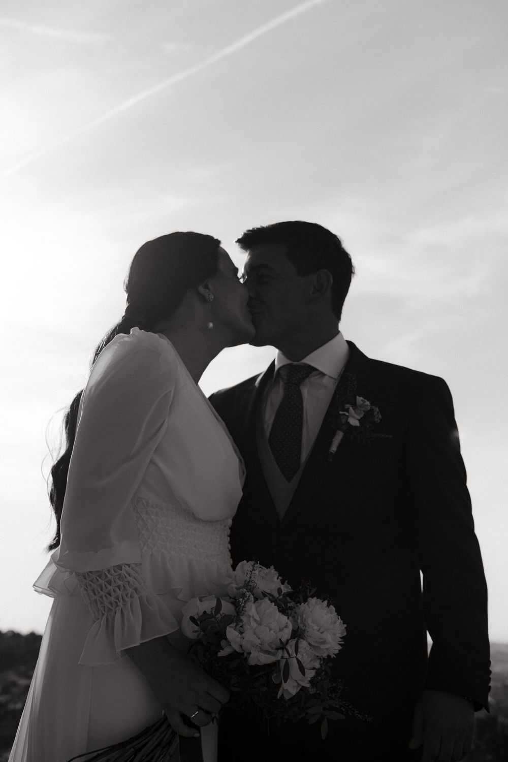 Black and white wedding photo of newlyweds sharing an intimate moment with a bouquet against a sunset sky.