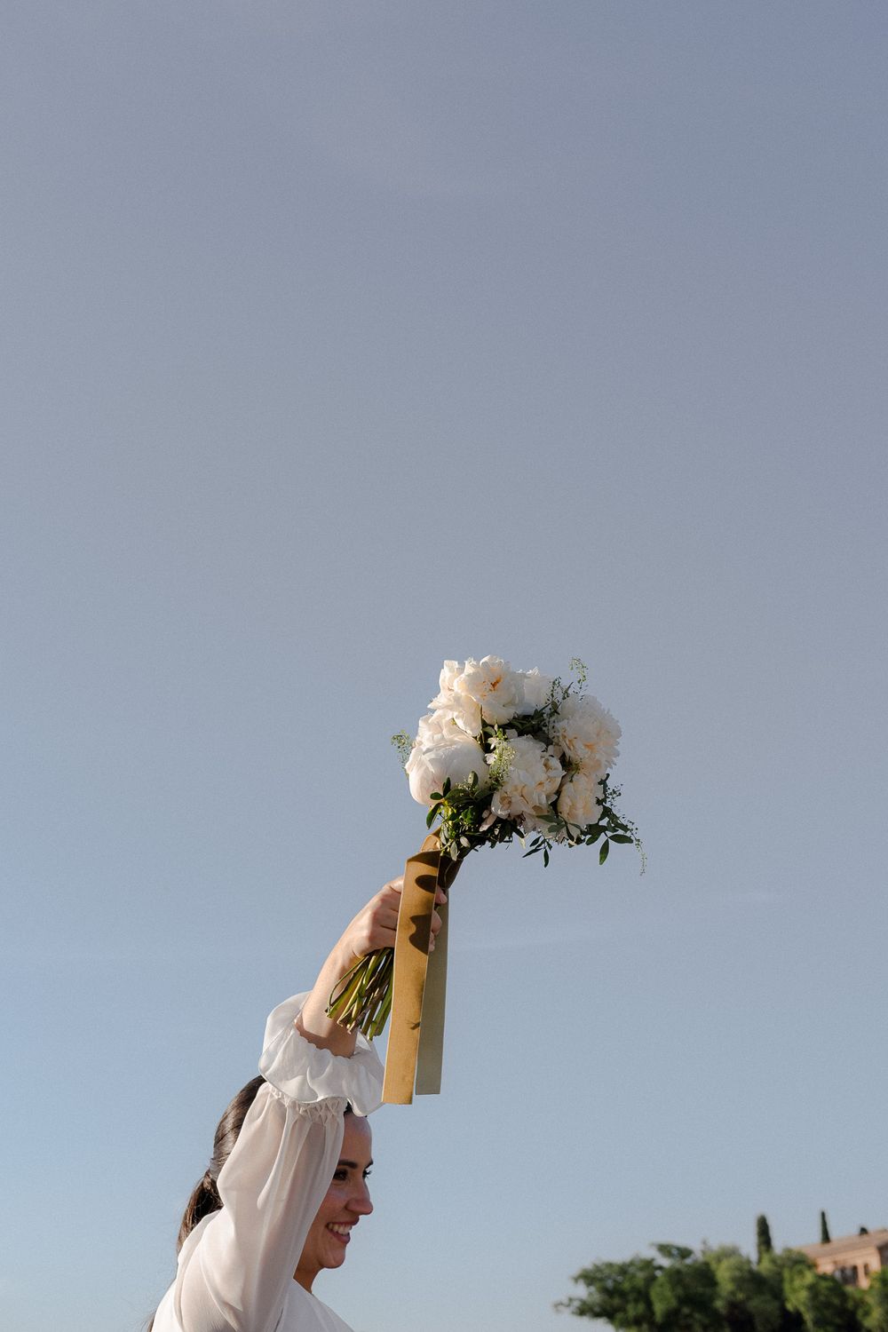 Joyful bride raising white bouquet against bright blue sky during celebration.