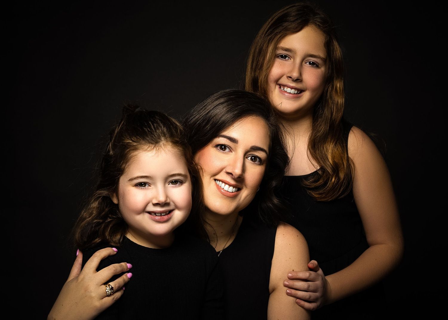 Three people in black clothing pose together against dark background in warm studio portrait.