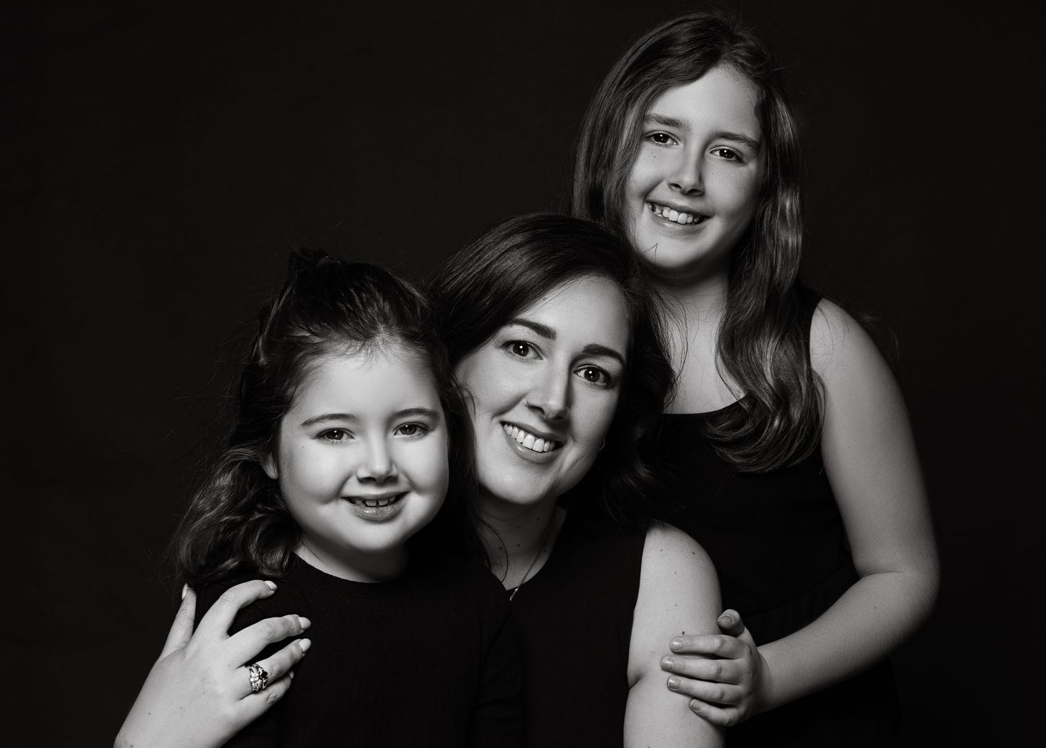 Black and white portrait of three smiling family members embracing against dark background with soft lighting.