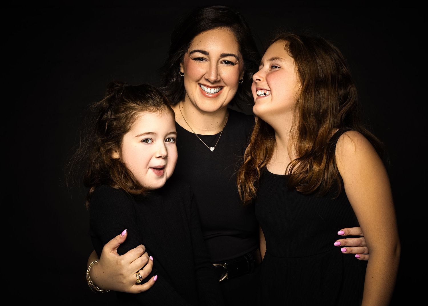 Three people wearing black outfits share a joyful moment together against a dark background.