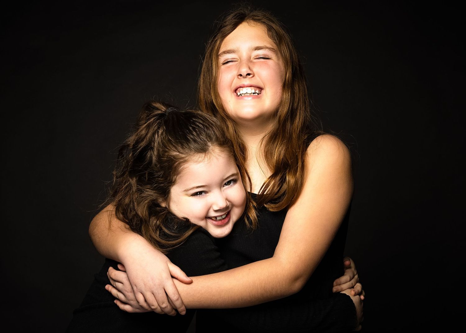 Two people embracing and laughing together in a candid studio portrait against a black background.