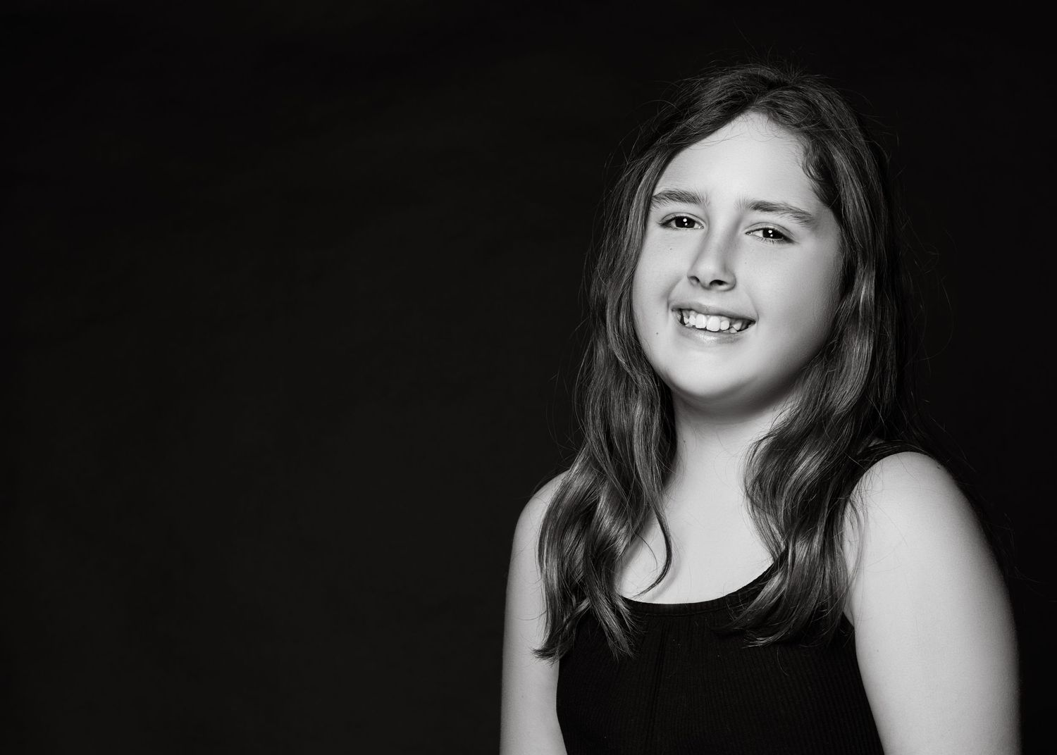 Black and white portrait of a teen smiling warmly wearing a dark sleeveless top against dark background.