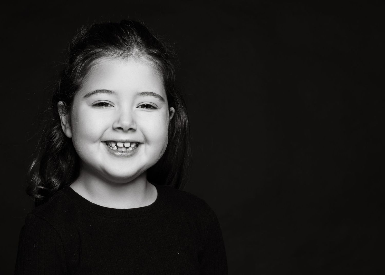Black and white studio portrait of a young child with an expressive smile wearing dark clothing.