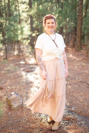 A person in a flowing cream dress and white top smiling while standing on a forest path.