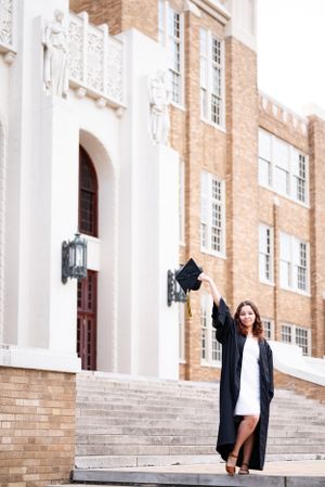 Graduate in cap and gown throwing mortarboard on steps of historic brick building with white trim.