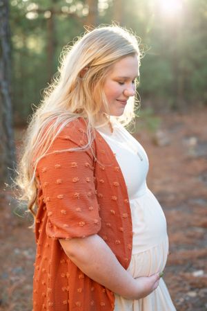 Glowing maternity portrait in a forest with golden sunlight and a rust-colored cardigan.