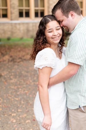 A couple embraces and laughs together on an autumn path near a stone building.