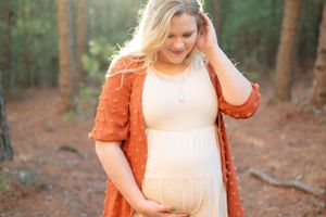 A maternity photo of someone in an orange cardigan touching their hair in dappled forest light.