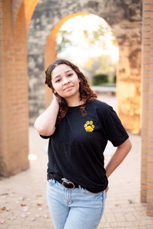 A casual portrait under brick archway wearing black t-shirt with paw print logo and jeans.