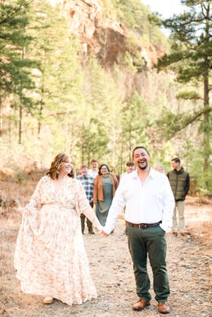 A couple laughing together on a forest path with others visible in the background.