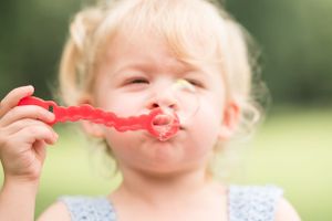 A toddler playfully eats a red fruit snack while playing outside.