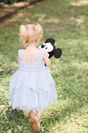 A child in a blue tulle dress holds a Mickey Mouse plush toy while walking in a garden.