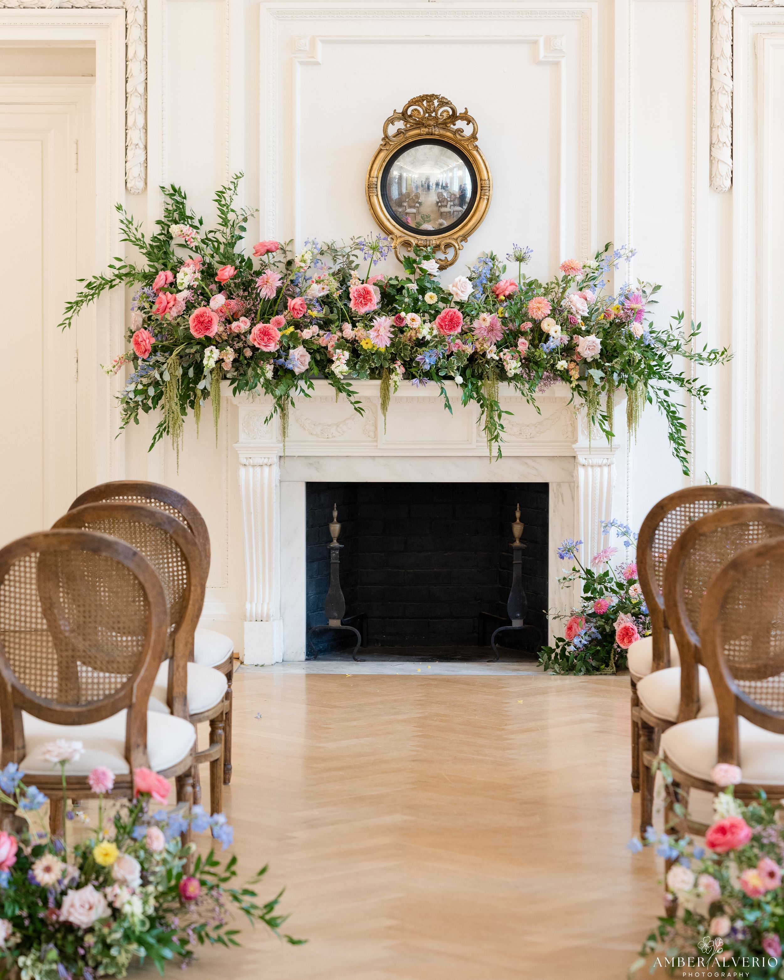 Ornate fireplace decorated with pink and white floral arrangements and vintage chairs for wedding ceremony.