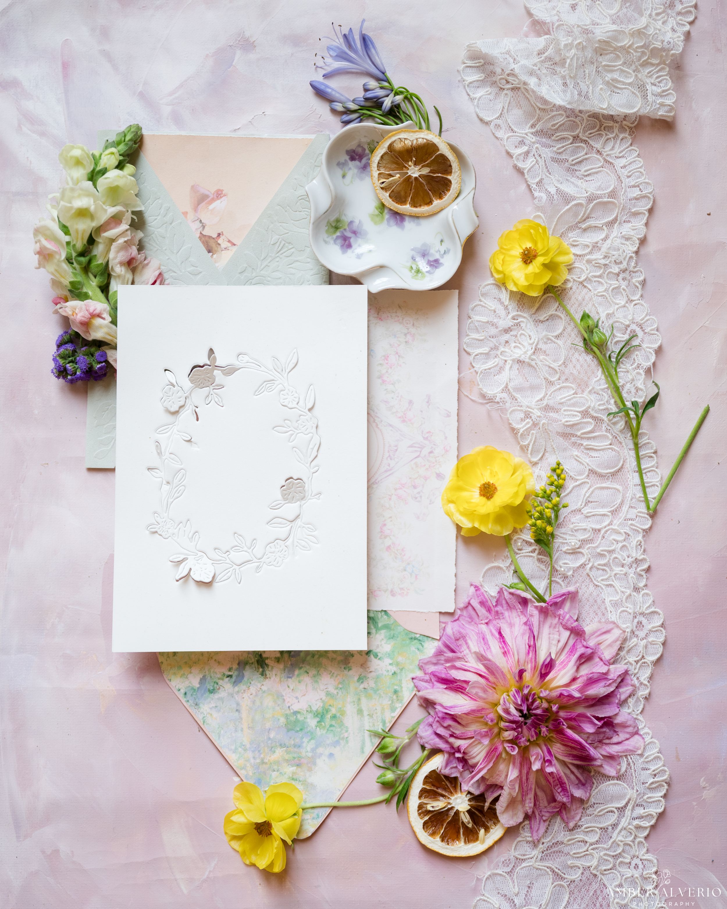 Styled wedding flatlay with pressed flowers, white stationery, dried citrus and delicate lace on pink background.