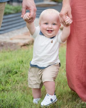 Happy toddler learning to walk while holding hands with parents on grassy lawn.