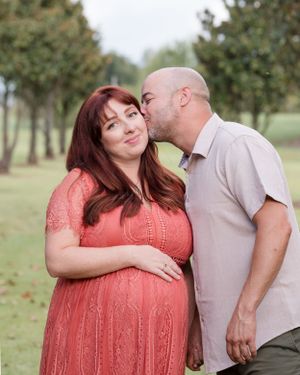 A couple shares a tender moment outdoors, with her wearing a coral lace dress and cradling her pregnant belly.