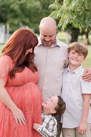 A joyful family portrait outdoors with a pregnant mother in a coral dress laughing together in a park setting.