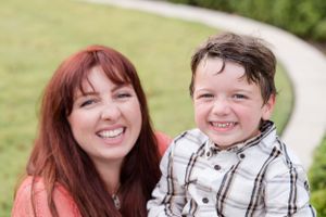 A parent and child smile together outdoors on a sunny day with green grass in the background.