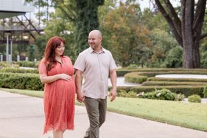A couple walks hand in hand along a garden path with green landscaping and trees in the background.