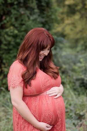 Expectant mother in coral lace dress tenderly holds her baby bump while standing in a garden setting.