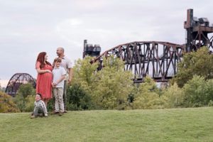 Two people and a small dog stand on grassy hill overlooking historic railroad bridge structure and green trees.