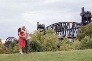Couple embracing on grassy hillside with industrial railway bridge and cloudy sky in the background.