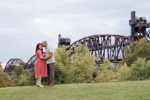 A couple embraces on a grassy hill with a rusted railroad bridge structure visible in the background.