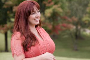 Woman in coral lace dress smiling outdoors with greenery in the background.