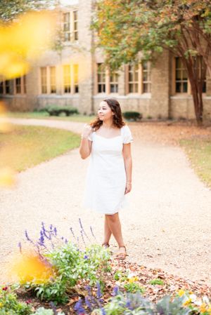A figure in a white dress stands on a path near purple flowers and autumn trees.