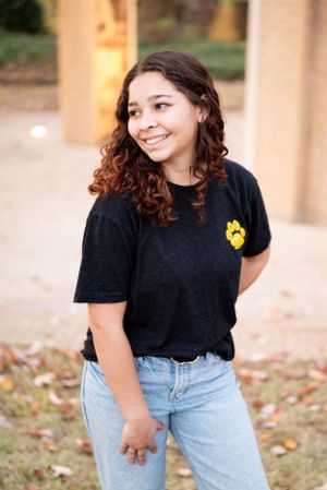 A person wearing a black t-shirt with a yellow paw print design and jeans poses outdoors near architectural columns.