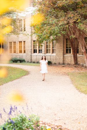 A person in a white dress walks along a stone path beside a historic brick building on a sunny autumn day.