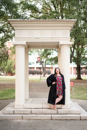 Graduate in black robe and floral dress poses under classical stone archway on campus.
