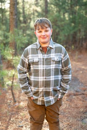 Someone in a plaid flannel shirt and brown pants standing casually on a forest trail.