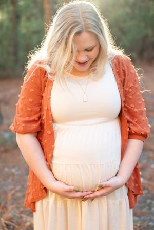 An expectant mother in a white dress and orange cardigan holding her belly in a forest setting.