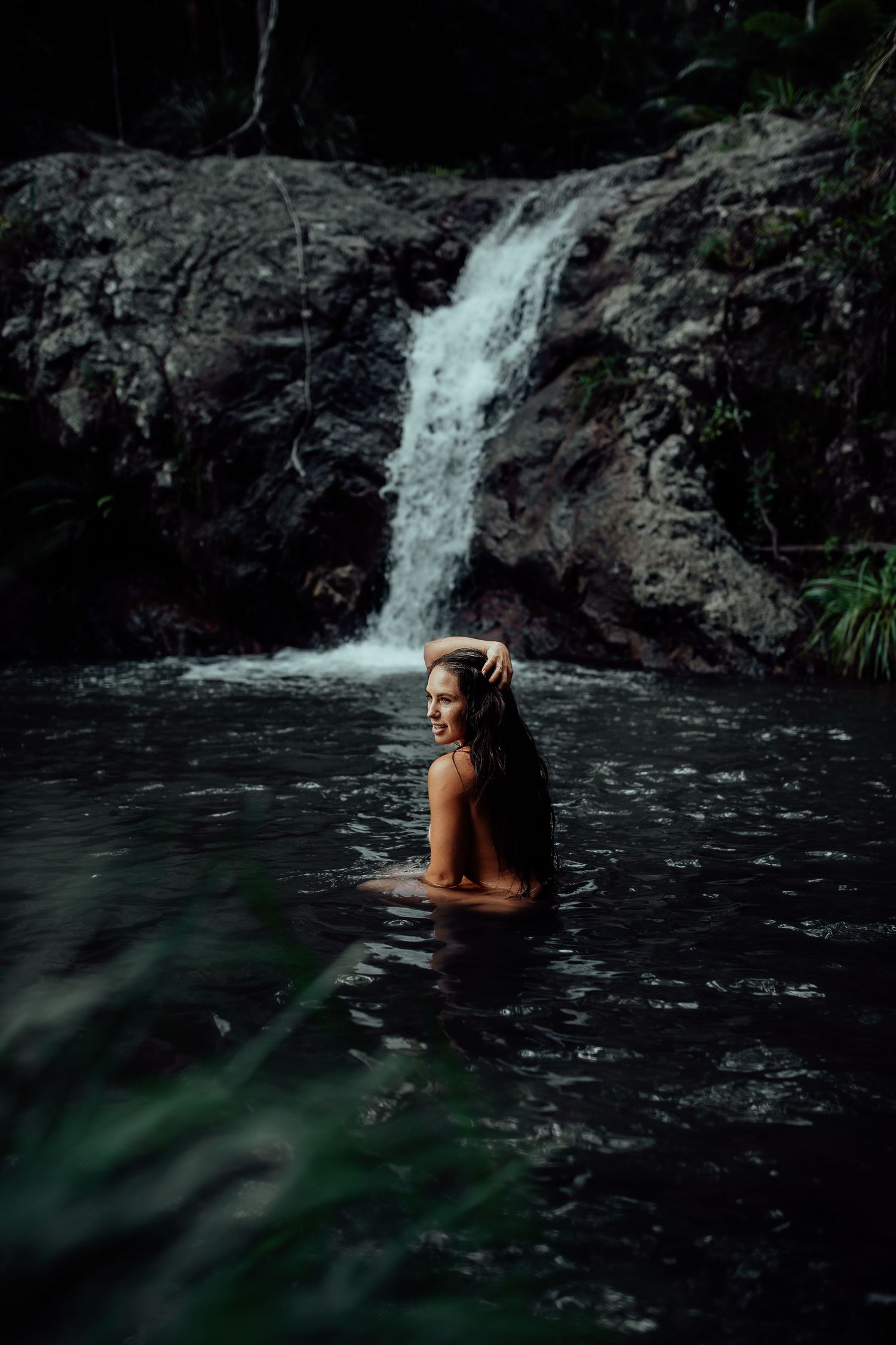 Swimming in dark waters beneath a cascading waterfall in a tropical forest setting.