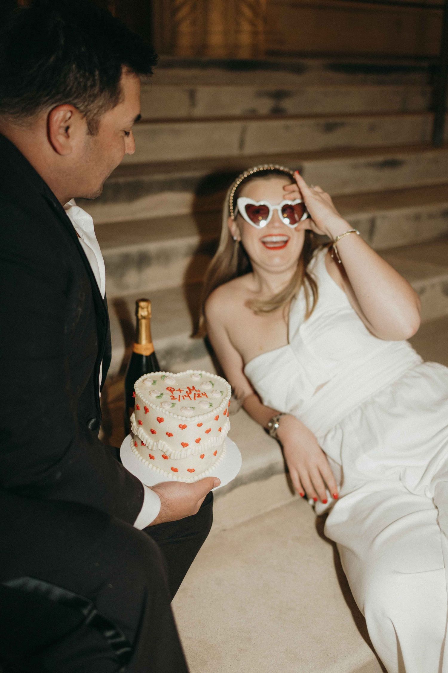 A couple shares a sweet moment with heart-decorated cake and champagne, the person in white wearing retro sunglasses.